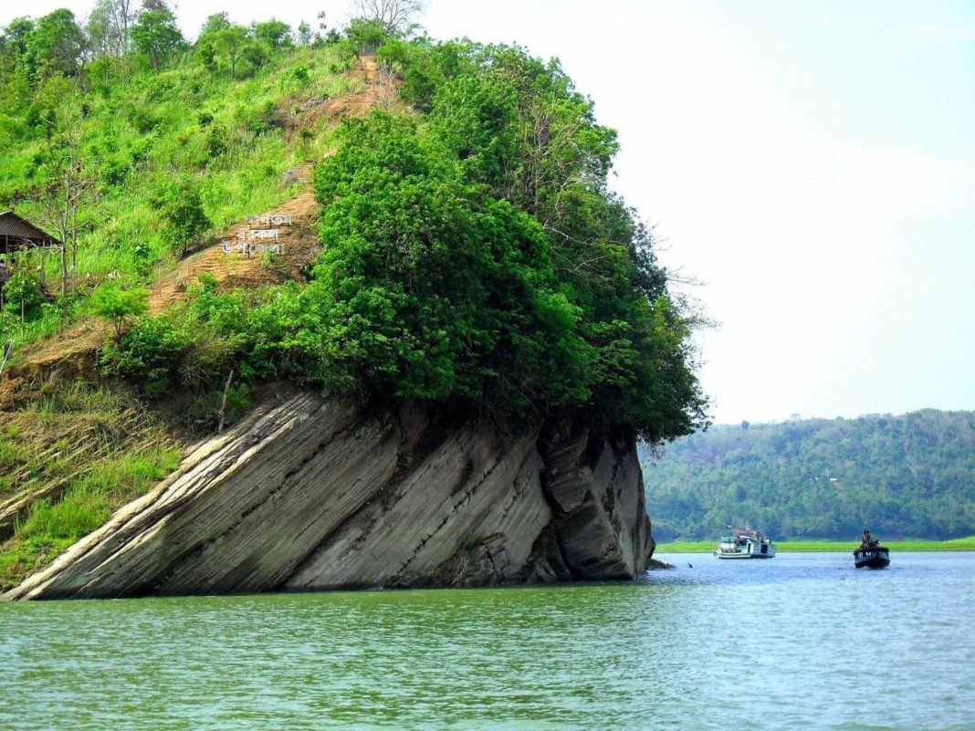 কাপ্তাই হ্রদের ধারে গাছে ঢাকা খাড়া পাথুরে পাহাড়, জলে কয়েকটি ছোট নৌকা