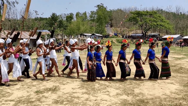 খোলা মাঠে সাদা মাথাবন্ধনী পরা ম্রো পুরুষেরা লম্বা নলের বাঁশের যন্ত্র হাতে সারি বেঁধে এগোচ্ছেন, পাশে নীল ব্লাউজ ও ফুলের মাথাসাজে হাত ধরে নাচছেন ম্রো নারীরা