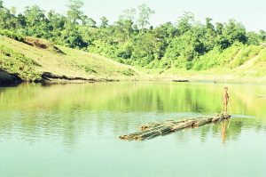 সবুজ পাহাড়ে ঘেরা শান্ত হ্রদের জলে বাঁশের ভেলায় দাঁড়িয়ে একজন, চারপাশে স্বচ্ছ জলের প্রতিফলন