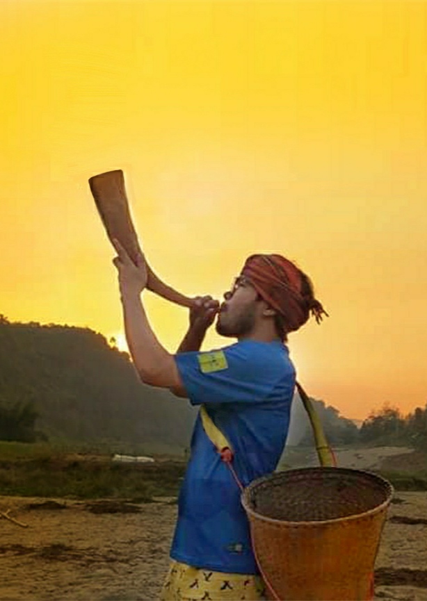 সূর্যাস্তের হলুদ আকাশের সামনে লাল মাথাবন্ধনী ও নীল জামা পরা এক তরুণ বাঁকা শিং মুখে তুলে ফুঁ দিচ্ছেন, কাঁধে বাঁশের ঝুড়ি