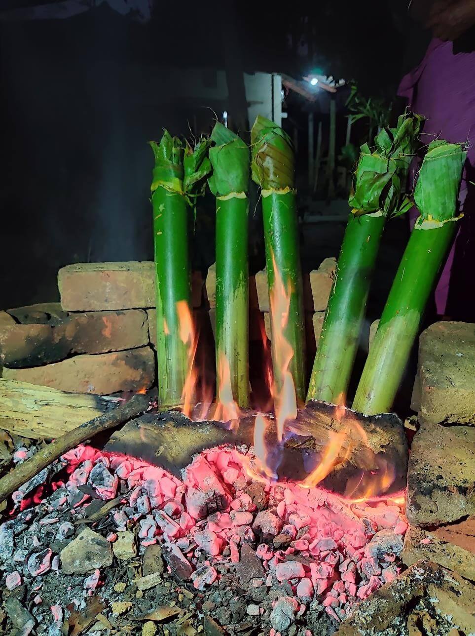 জ্বলন্ত কয়লার ওপর খাড়া করে রাখা পাঁচটি সবুজ বাঁশের চোঙ, প্রতিটির মুখ সবুজ পাতা দিয়ে বন্ধ, নিচে আগুনের শিখা