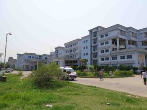 সাদা রঙের বহুতল হাসপাতাল ভবনের সামনে খোলা চত্বর, পাশে অ্যাম্বুলেন্স ও গাছপালা