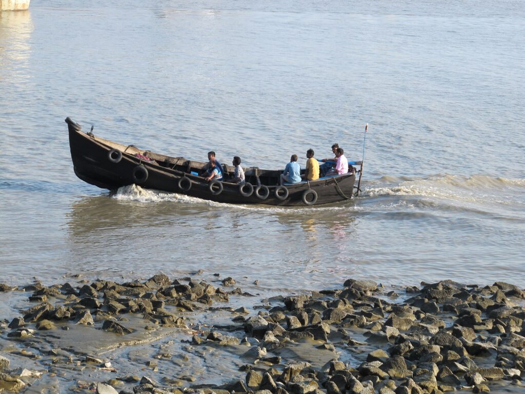 ঘোলা জলের কর্ণফুলী নদীতে কাঠের লম্বা নৌকা, তাতে কয়েকজন যাত্রী; সামনের তীরে ছড়ানো পাথর