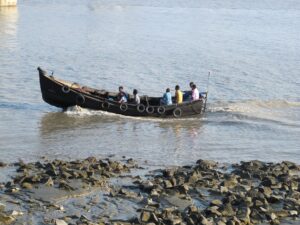 ঘোলা জলের কর্ণফুলী নদীতে কাঠের লম্বা নৌকা, তাতে কয়েকজন যাত্রী; সামনের তীরে ছড়ানো পাথর