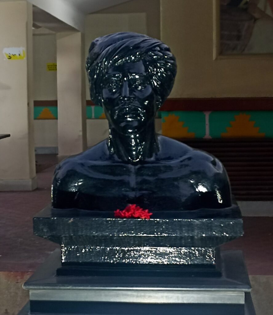 কালো পাথরের বেদির ওপর বিরসা মুন্ডার আবক্ষ ভাস্কর্য, গলায় লাল ফুলের মালা; পিছনে রঙিন দেয়াল