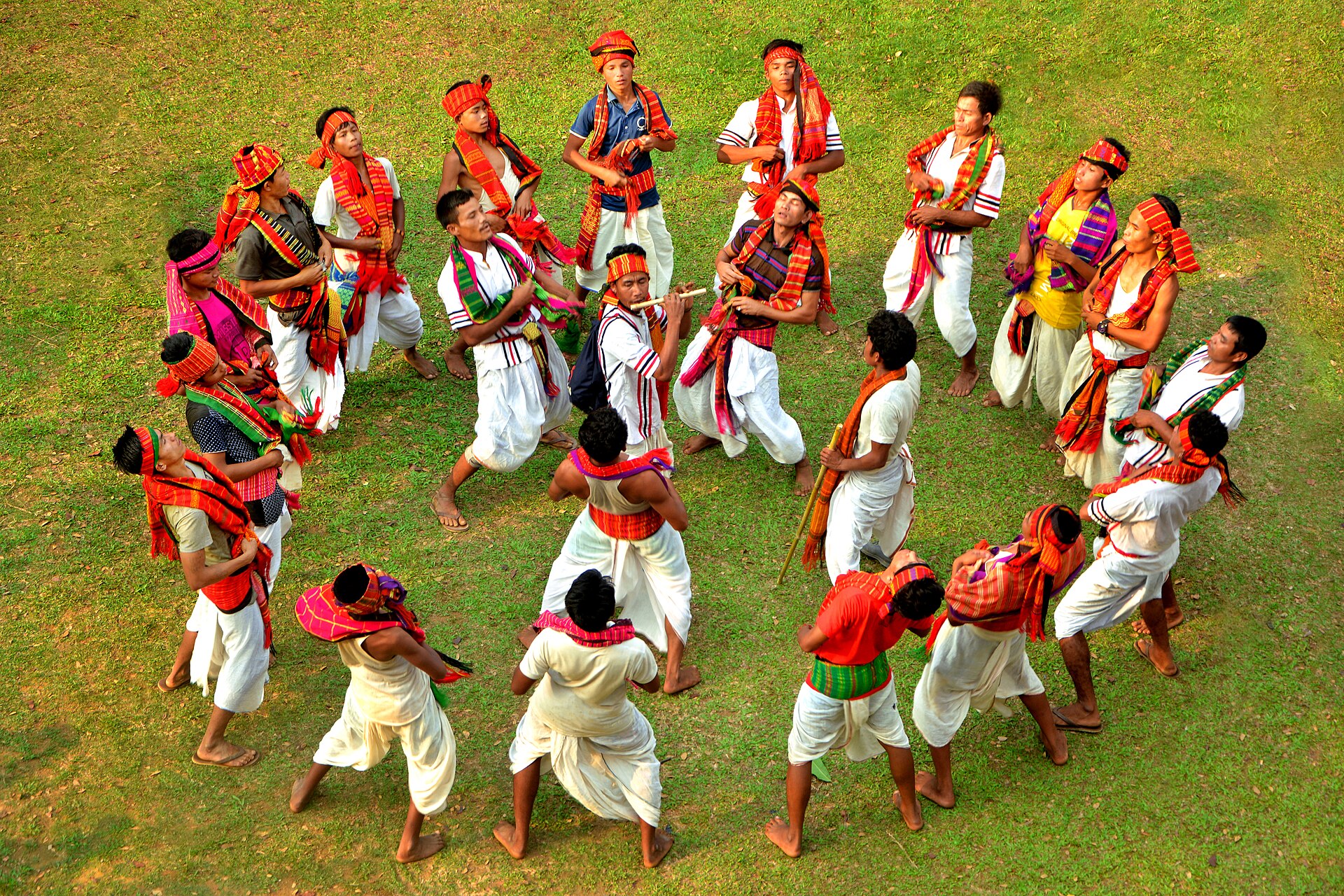সবুজ মাঠে গোল হয়ে দাঁড়িয়ে লাল-কমলা নকশার উত্তরীয় ও পাগড়ি পরা ত্রিপুরা নৃত্যশিল্পীরা গরাইয়া নৃত্য করছেন, মাঝের একজনের হাতে বাঁশি