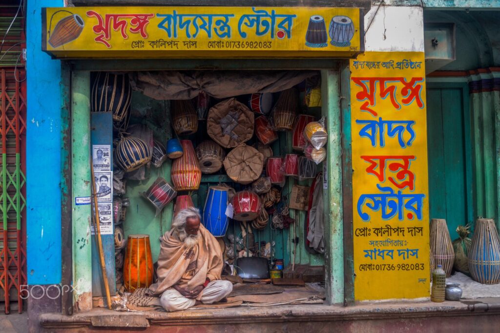 রঙিন দোকানের সামনে থরে থরে সাজানো ঢোল, মৃদঙ্গ ও অন্যান্য বাদ্যযন্ত্র; দোকানের সাইনবোর্ডে বাংলায় লেখা মৃদঙ্গ বাদ্যযন্ত্র স্টোর