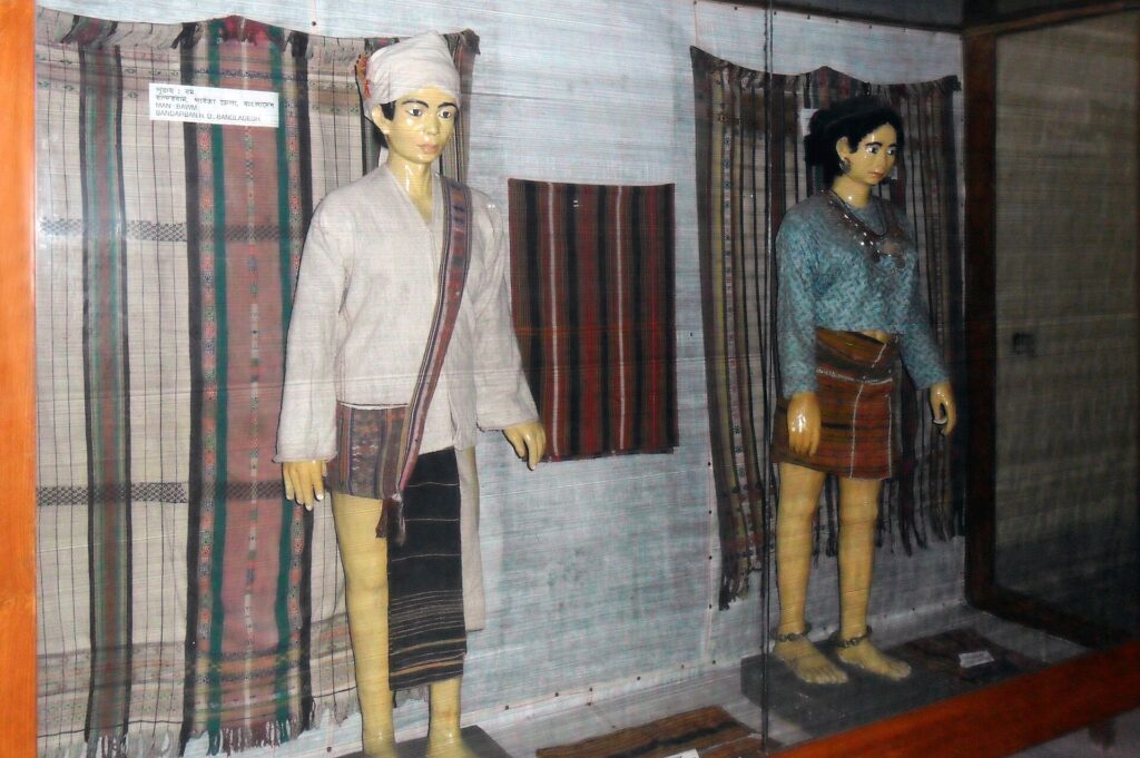 জাদুঘরের কাচের প্রদর্শনীতে ঐতিহ্যবাহী পোশাক পরা বম পুরুষ ও নারীর দুটি মূর্তি, পেছনে ঝোলানো ডোরাকাটা বোনা কাপড়