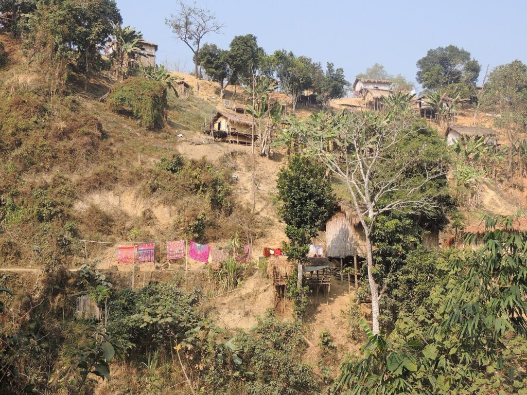 Thatched and tin-roofed houses on stilts scattered across a dry, terraced hillside, with colourful cloths drying on a line and banana plants in the foreground