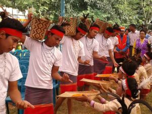 লাল কপালবন্ধনী, সাদা গেঞ্জি ও লাল পাড়ের লুঙ্গি পরা তরুণেরা কাঁধে বেতের ঝুড়ি নিয়ে সারি বেঁধে নাচছেন, সামনে পালক-গোঁজা লাল ফিতা পরা মেয়েরা কুলায় গাঁদা ফুল নিয়ে বসে