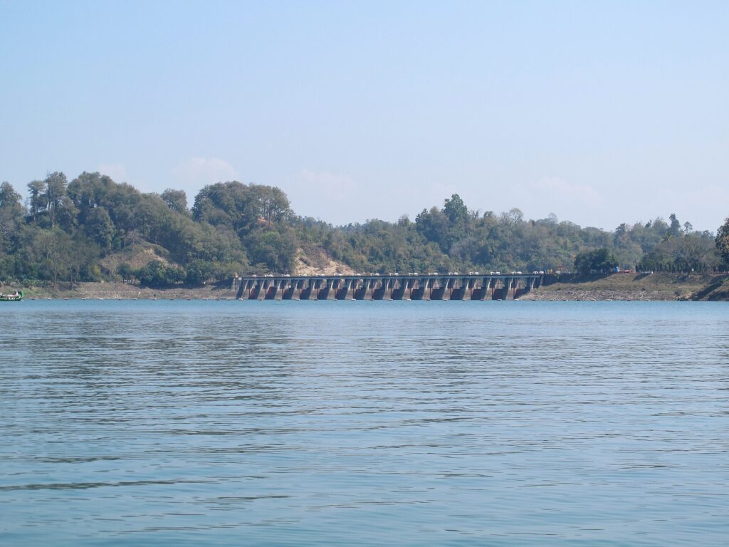 কাপ্তাই হ্রদের জলের ওপারে কাপ্তাই বাঁধের সারি সারি স্লুইস গেট, পেছনে গাছে ঢাকা টিলা ও পরিষ্কার নীল আকাশ