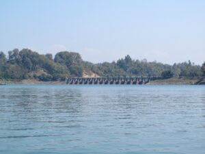 কাপ্তাই হ্রদের জলের ওপারে কাপ্তাই বাঁধের সারি সারি স্লুইস গেট, পেছনে গাছে ঢাকা টিলা ও পরিষ্কার নীল আকাশ