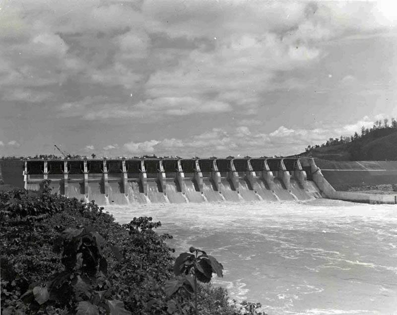 সাদাকালো ছবিতে কাপ্তাই বাঁধের স্পিলওয়ে, সারি সারি জলকপাট দিয়ে সাদা ফেনা তুলে পানি নামছে, সামনে ঝোপঝাড়