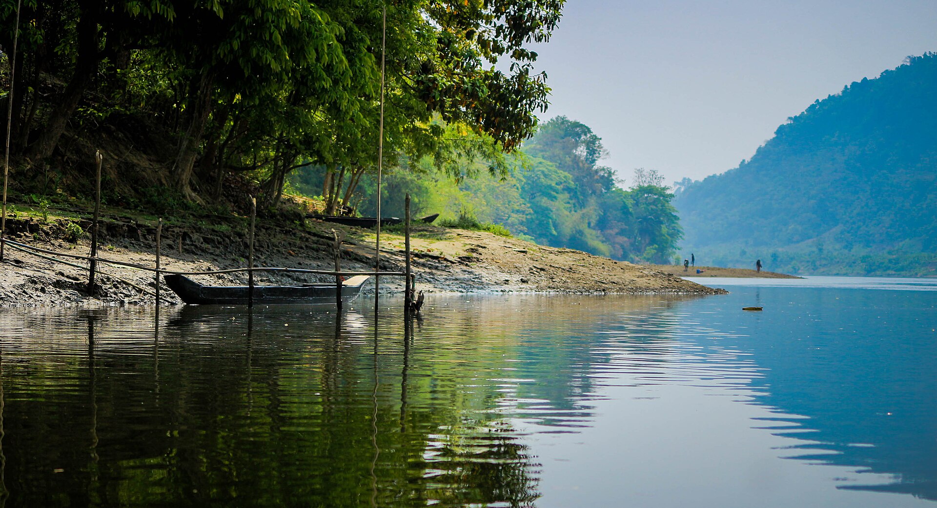 কাপ্তাই হ্রদের কিনারে পানি নেমে যাওয়ায় খোলা কাদামাটির পাড়, খুঁটিতে বাঁধা একটি কাঠের নৌকা আর দূরে পাড়ে দাঁড়ানো কয়েকজন মানুষ