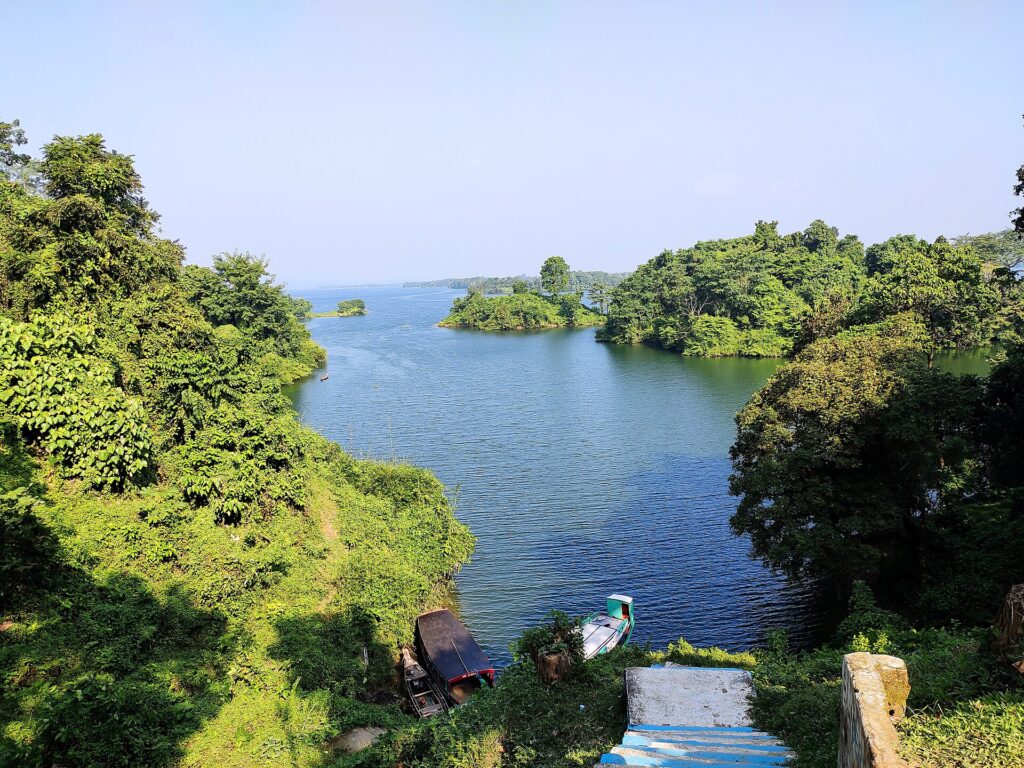 Kaptai Lake seen from a green hillside, with forested islands and two boats moored at the foot of steps