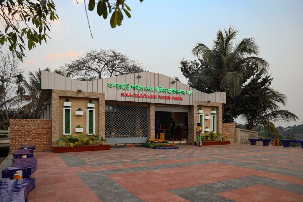 খাগড়াছড়ি পার্বত্য জেলা পরিষদ হর্টিকালচার পার্কের প্রবেশ-ভবন, সামনে টালি-বাঁধানো চত্বর ও নারকেল গাছ, ভবনের মাথায় বাংলা ও ইংরেজিতে পার্কের নাম লেখা