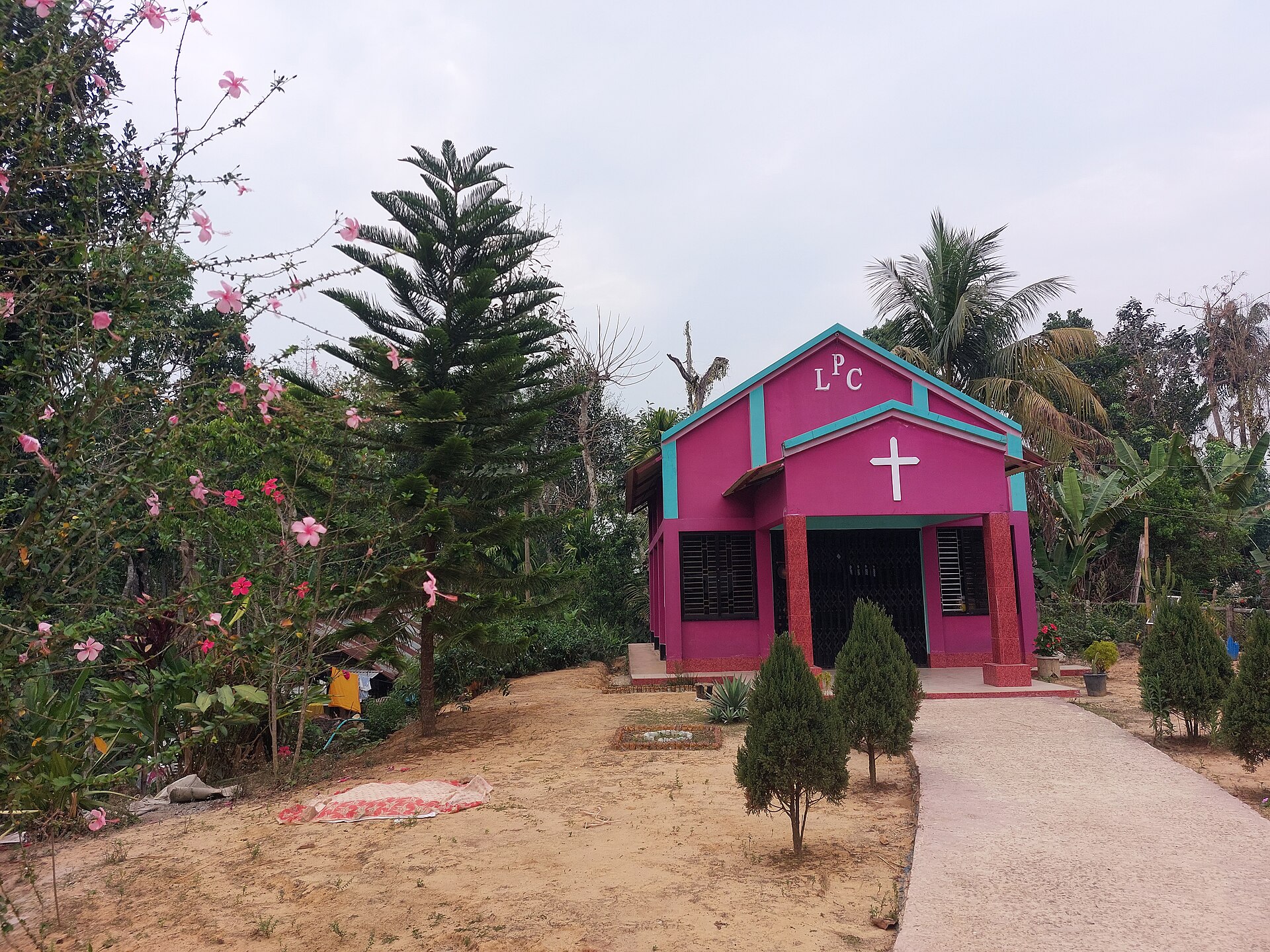 গোলাপি দেয়াল ও সবুজ কিনারার ছোট একটি গির্জা, সামনে ক্রুশচিহ্ন, পাশে ঝাউ, নারকেল ও কলাগাছ