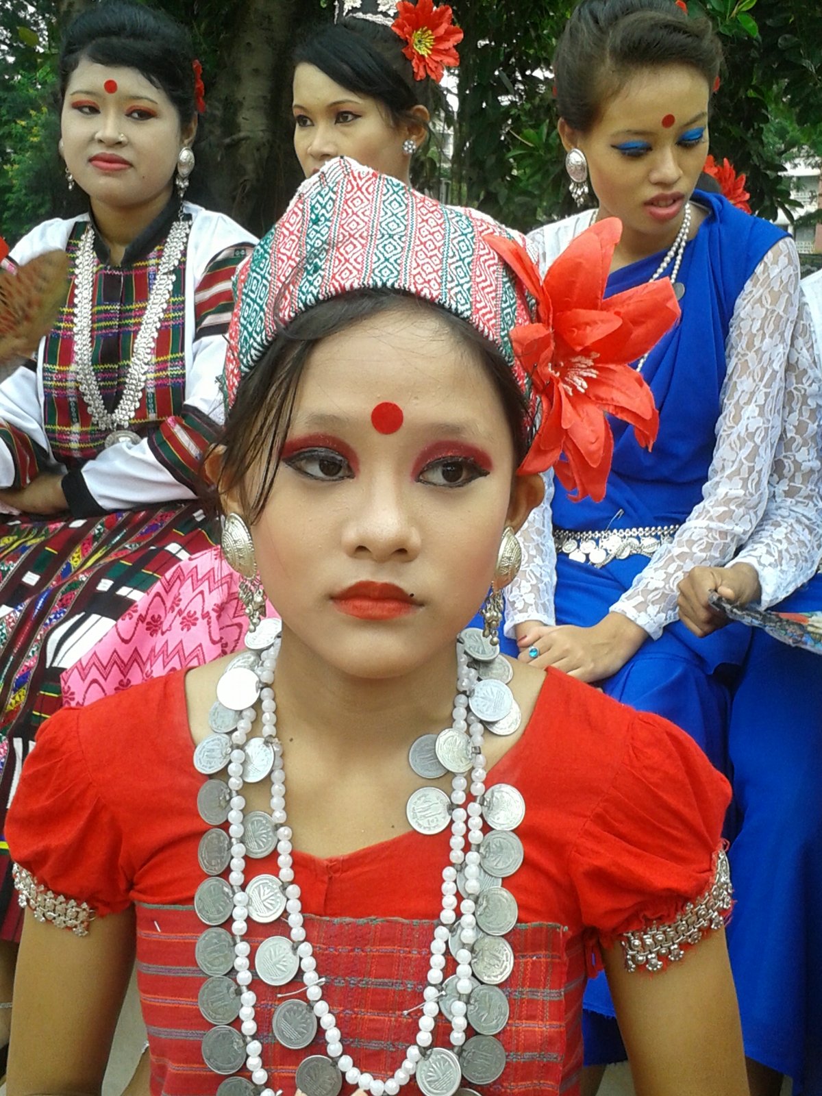 লাল ব্লাউজ, নকশা-করা মাথার কাপড়, চুলে লাল ফুল আর গলায় রুপার মুদ্রা ও পুঁতির মালা পরা খুমি তরুণী, পেছনে বসে আছেন আরও কয়েকজন