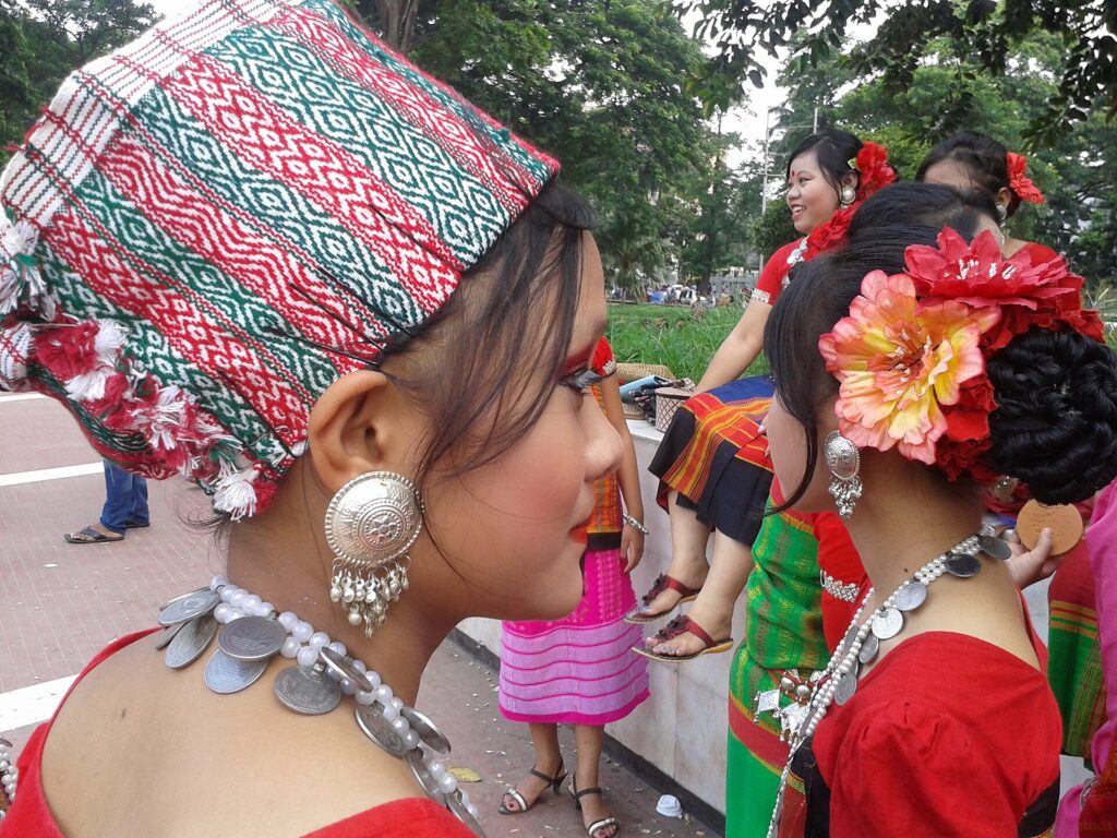 ঐতিহ্যবাহী নকশা-করা মাথার কাপড়, রুপার বড় কানের দুল ও মুদ্রার মালা পরা খুমি তরুণী, পেছনে লাল পোশাকে আরও কয়েকজন নৃত্যশিল্পী