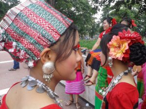 ঐতিহ্যবাহী নকশা-করা মাথার কাপড়, রুপার বড় কানের দুল ও মুদ্রার মালা পরা খুমি তরুণী, পেছনে লাল পোশাকে আরও কয়েকজন নৃত্যশিল্পী