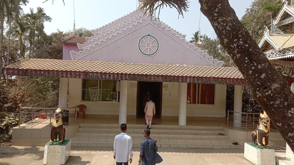কুয়াকাটার একটি বৌদ্ধ মন্দিরের সম্মুখভাগ: টালির ছাদের বারান্দা, সাদা স্তম্ভ, চূড়ায় ধর্মচক্র, সিঁড়ির দুই পাশে দুটি সিংহমূর্তি