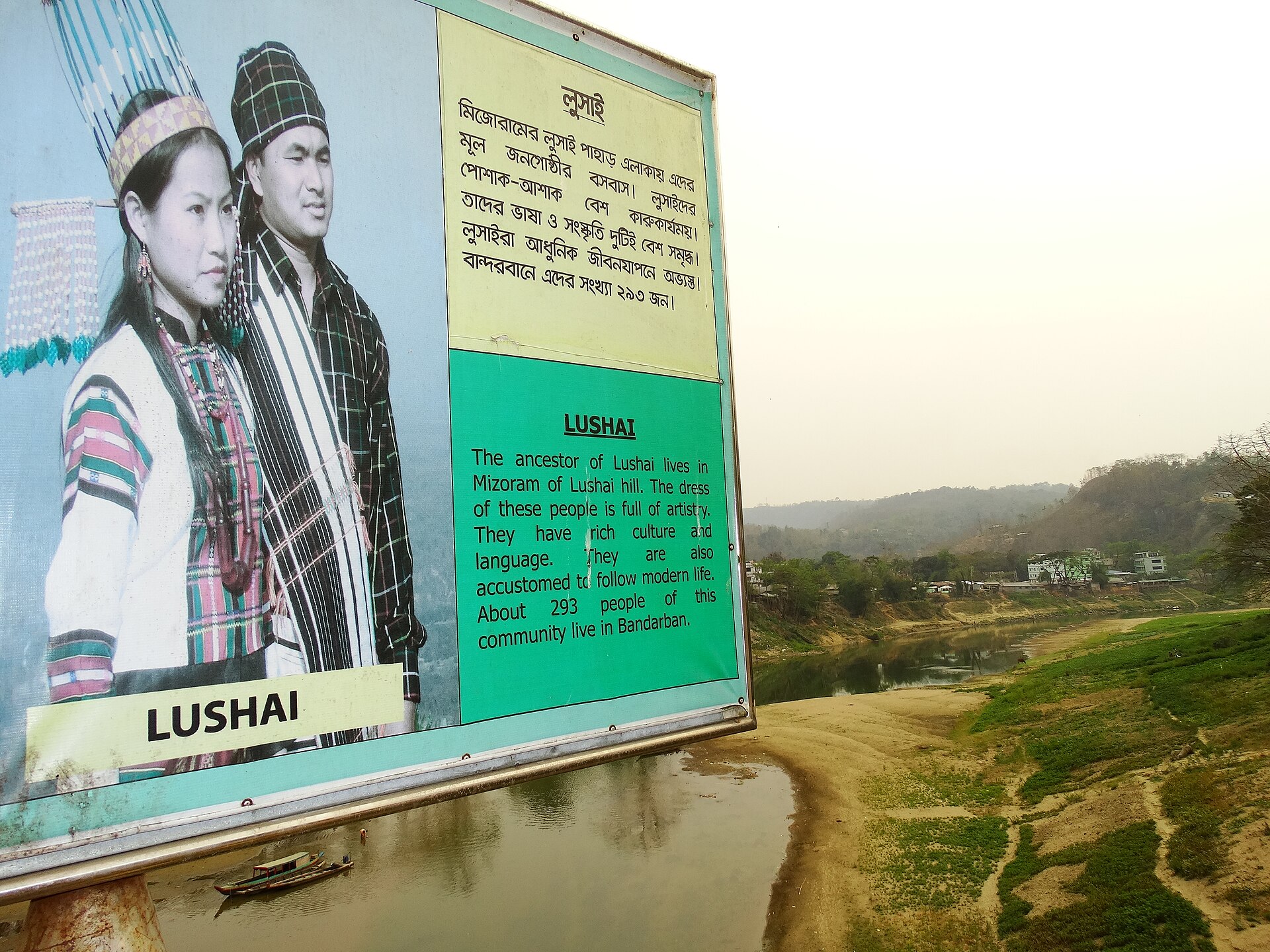বান্দরবানে সাঙ্গু নদীর সেতুর পাশে বাংলা ও ইংরেজিতে লেখা লুসাই পরিচিতি ফলক, বাঁ পাশে ঐতিহ্যবাহী পোশাকে দুজনের ছবি, পেছনে নদী ও পাহাড়
