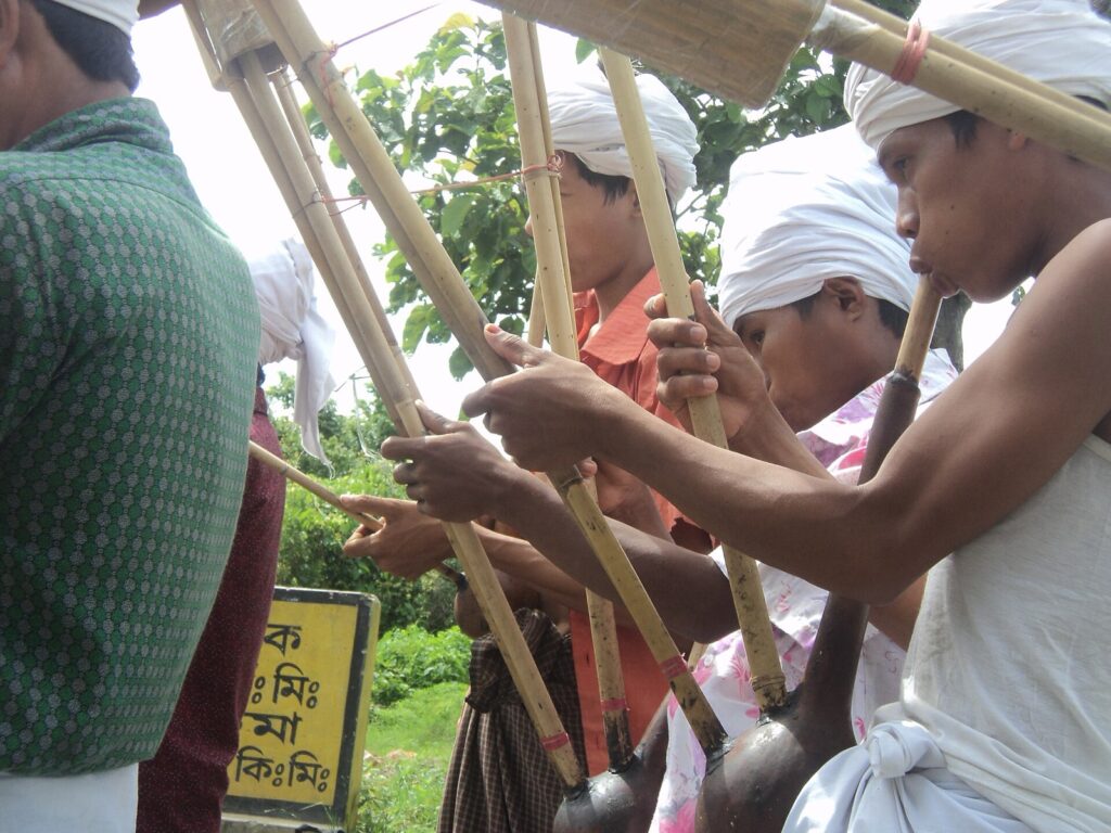 সাদা পাগড়ি পরা কয়েকজন ম্রো তরুণ লাউয়ের খোলে বসানো লম্বা বাঁশের নলের প্লুং মুখে ধরে বাজাচ্ছেন, নলের মাথায় বাঁশের ঢাকনা