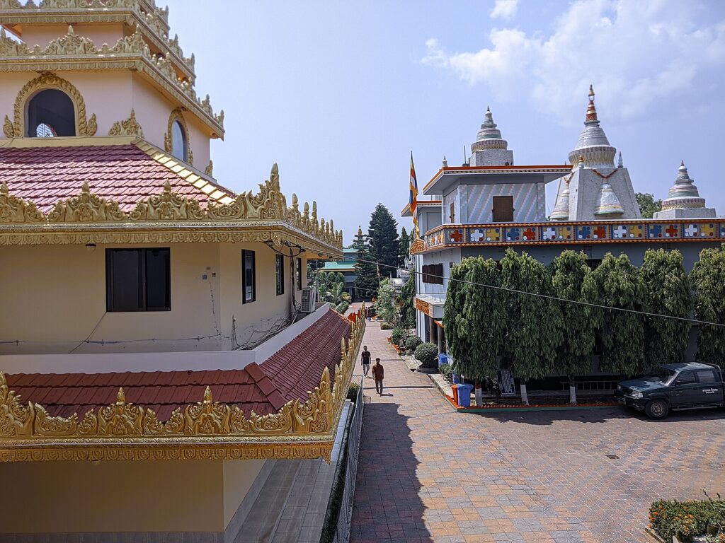 রাঙামাটির রাজবন বিহার প্রাঙ্গণ: সোনালি কারুকাজ করা ছাদের ভবন, ডান পাশে চূড়াওয়ালা বিহার ভবন ও পতাকা, মাঝখানে ইট বিছানো পথ