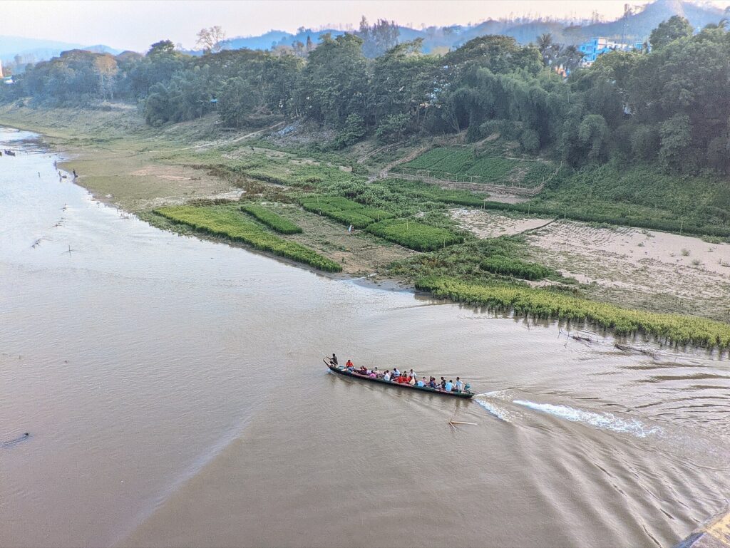 ওপর থেকে তোলা বান্দরবানে সাঙ্গু নদীর বাদামি জল, যাত্রী-ভরা একটি কাঠের নৌকা চলছে; পাড়ে সবুজ চাষের জমি, গাছপালা আর দূরে পাহাড়