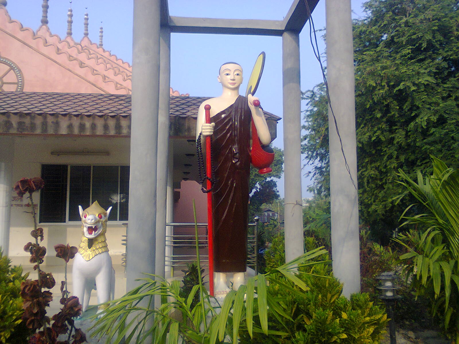 কুয়াকাটার সীমা বৌদ্ধ মন্দির প্রাঙ্গণে খয়েরি চীবর পরা ভিক্ষুর দাঁড়ানো মূর্তি, পাশে সাদা সিংহমূর্তি, পেছনে মন্দিরের টালির ছাদ