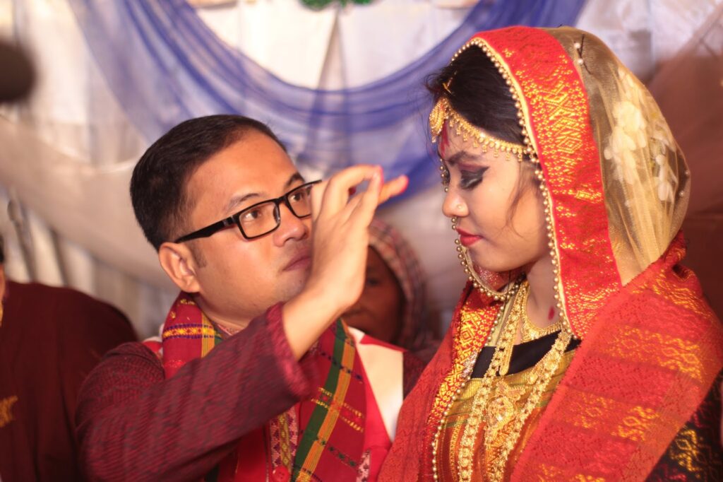 বিয়ের অনুষ্ঠানে লাল-সোনালি পোশাক ও গয়না পরা কনের কপালে হাত দিয়ে কিছু পরিয়ে দিচ্ছেন চশমা পরা বর, পেছনে নীল-সাদা কাপড়ের সাজ
