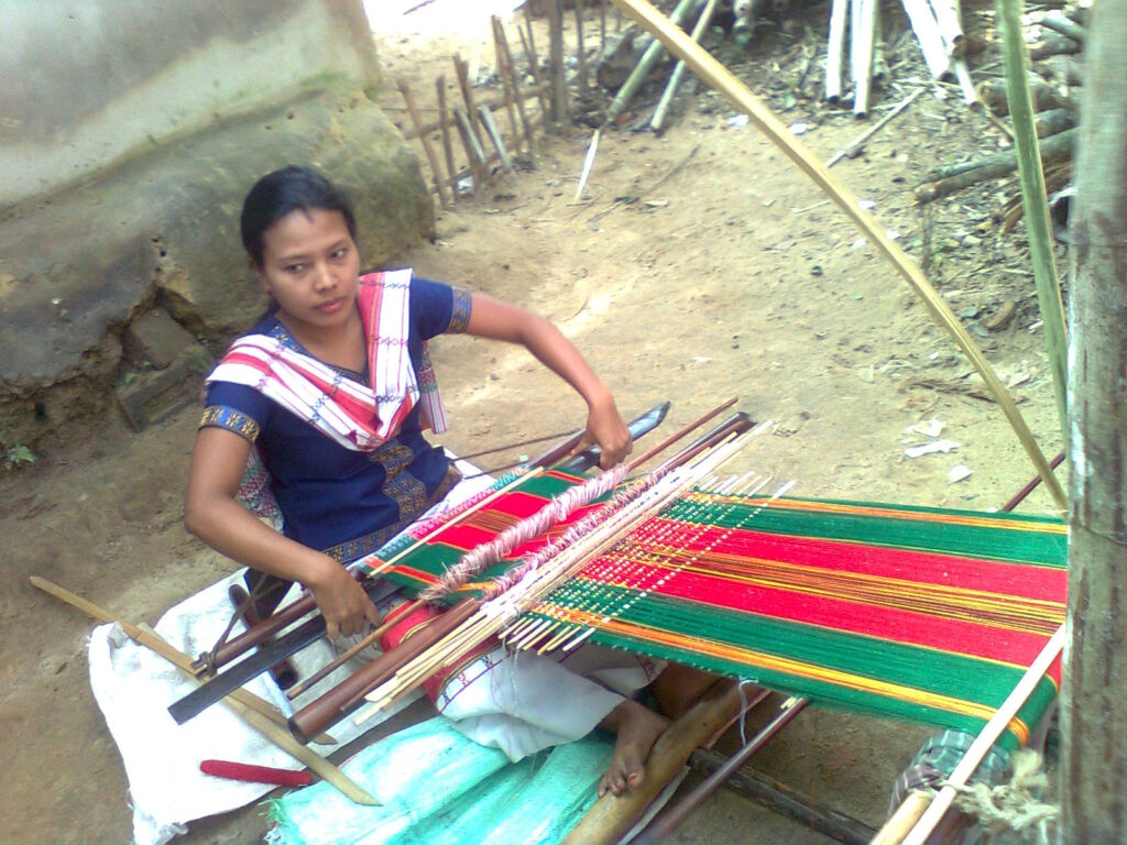 উঠানে মাটিতে বসে কোমর তাঁতে সবুজ-লাল ডোরাকাটা কাপড় বুনছেন একজন ত্রিপুরা নারী, কাঁধে সাদা-লাল নকশার কাপড়