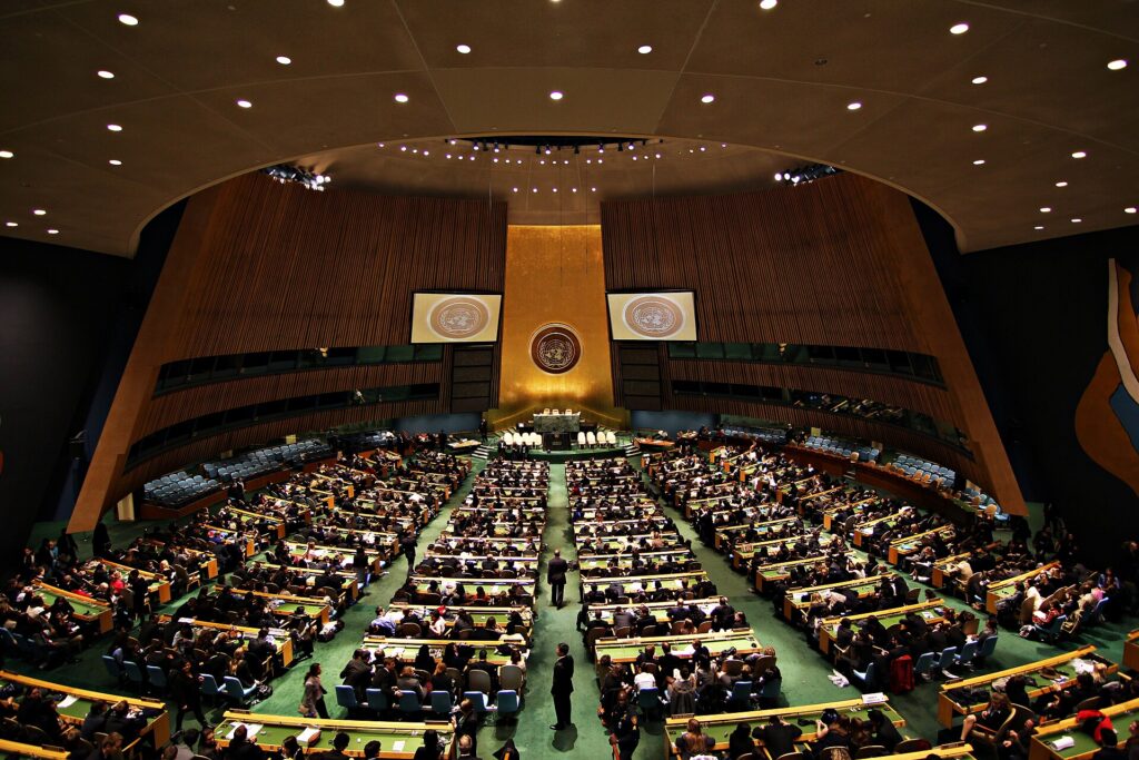 The United Nations General Assembly Hall in New York, with delegates seated in rows of desks facing the podium and the UN emblem on the gold wall