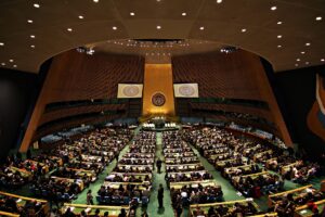 The United Nations General Assembly Hall in New York, with delegates seated in rows of desks facing the podium and the UN emblem on the gold wall